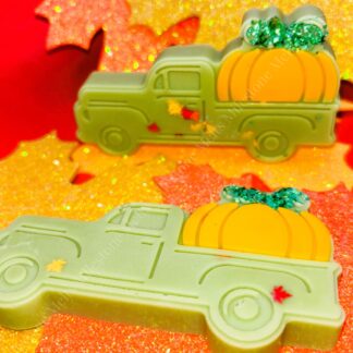 Autumn-themed wax melts shaped like vintage pickup trucks carrying bright orange pumpkins, decorated with glitter and small fall leaves, displayed on a vibrant background of sparkly red, orange, and yellow autumn leaves