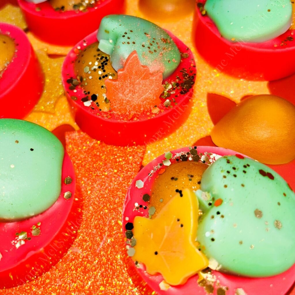 Colourful handmade autumn wax melts with apple, pear, and leaf designs, featuring vibrant red, orange, and green tones, sprinkled with glitter and decorative specks, displayed on a shimmering golden background.