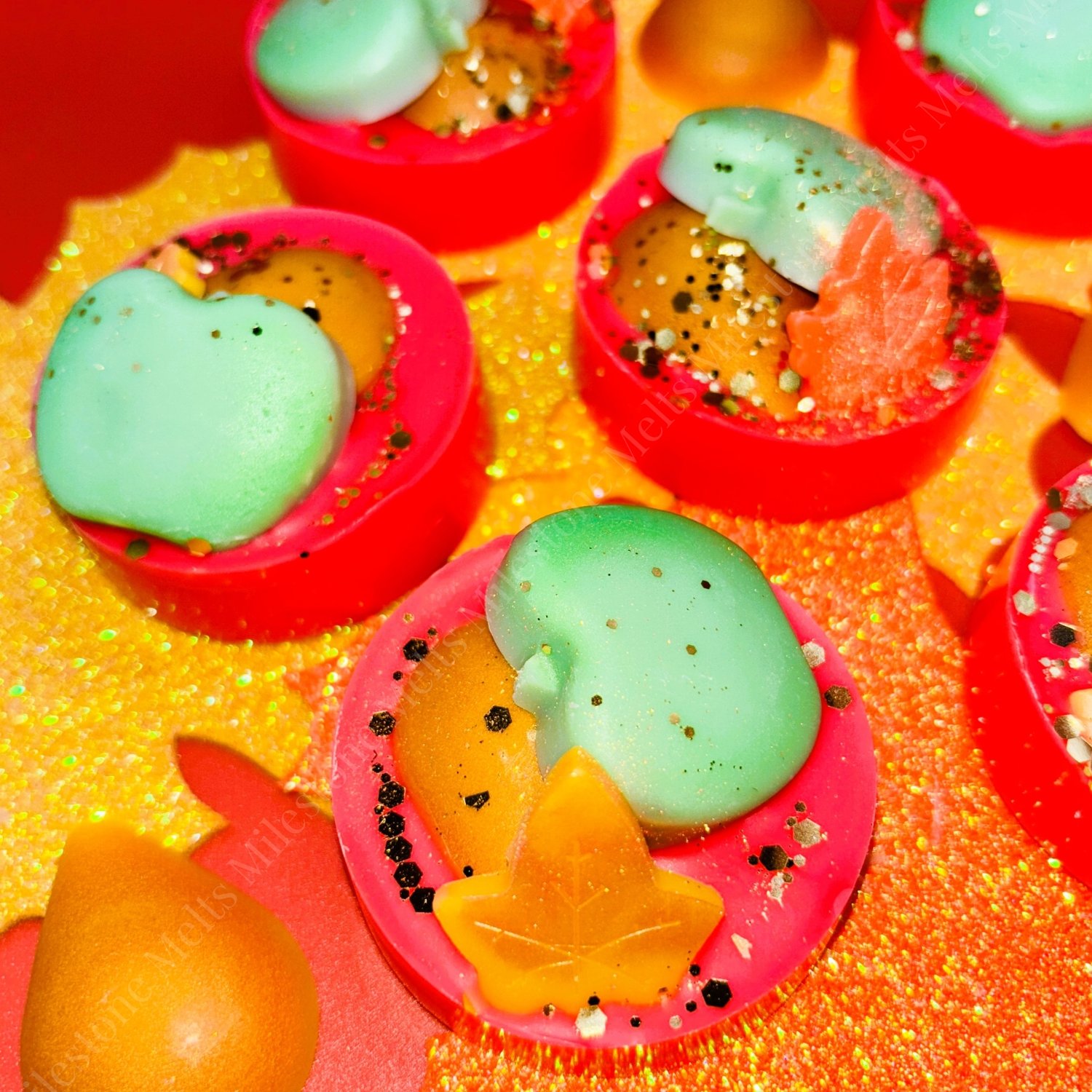 Golden Pear Harvest Wax Melt 2 Colourful handmade autumn wax melts with apple, pear, and leaf designs, featuring vibrant red, orange, and green tones, sprinkled with glitter and decorative specks, displayed on a shimmering golden background.